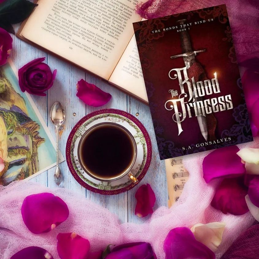 The Blood Princess book cover amongst a cup of tea, a book and pink flower petals.